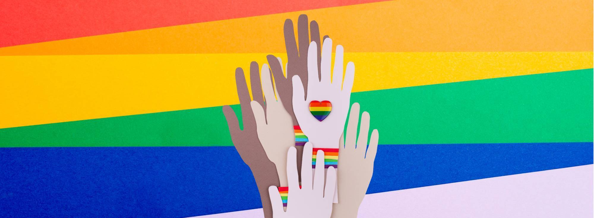 Image with rainbow colors that are symbolic of PRIDE Month celebrations. Uplifted hands of all races with rainbow heart images on the hands. 